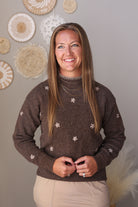 A smiling woman with long light brown hair stands indoors wearing a brown knit sweater with small white floral motifs and beige skirt. The background is a warm gray wall decorated with circular woven wall art.