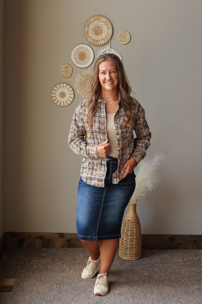 A woman stands indoors modeling a long-sleeve plaid button-up shirt in tan, brown, and blue tones, paired with a beige top, a blue denim skirt, and white sneakers. The background shows a neutral beige wall with decorative woven wall art and a wicker vase with dried grass.