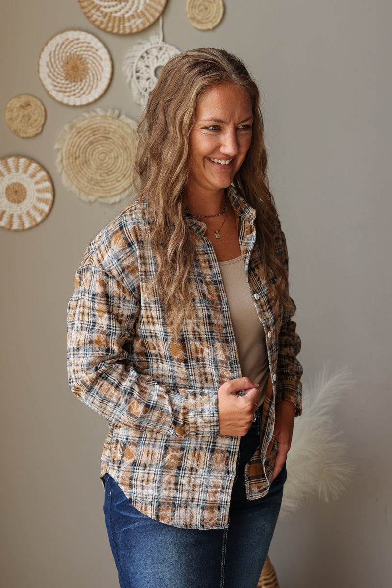 A woman wearing a tan and brown plaid flannel shirt over a beige top and blue jean skirt, standing against a neutral wall decorated with woven circular wall art.