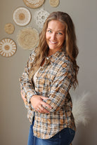 A smiling woman with long light brown hair stands indoors wearing a tan and brown plaid button-up shirt over a white top and blue jean skirt. The background is a warm gray wall with woven decorative wall hangings and a white feather prop.