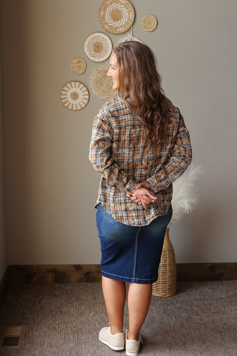 A woman stands with her back to the camera wearing a brown and beige plaid button-down shirt and a blue denim skirt in a styled room with neutral walls and decorative woven wall art.