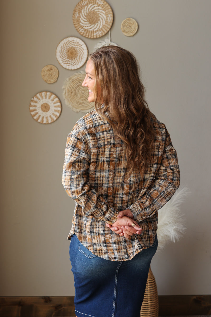A woman with long wavy brown hair is shown from the back wearing a brown and beige plaid flannel shirt and blue jean skirt, standing in front of a neutral wall decorated with woven wall art.