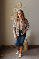 A woman stands indoors wearing a beige and brown plaid button-up shirt over a cream top, a dark blue denim skirt, and off-white sneakers. She is posed in front of a beige wall decorated with woven circular wall art and a tall woven vase with dried pampas grass.