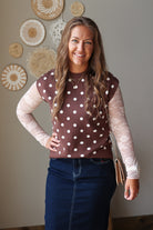 A woman modeling a brown mock-neck top with white polka dots over a sheer beige lace long-sleeve shirt, paired with a dark blue denim skirt.