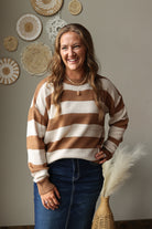 A woman smiling and wearing a brown and cream striped knit sweater with dark blue jean skirt, standing in a styled room with woven wall decor and a vase of pampas grass.