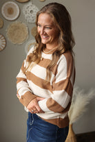 A woman wearing a brown and cream waffle-knit striped sweater and blue jean skirt, standing in a room with boho-style wall decor.