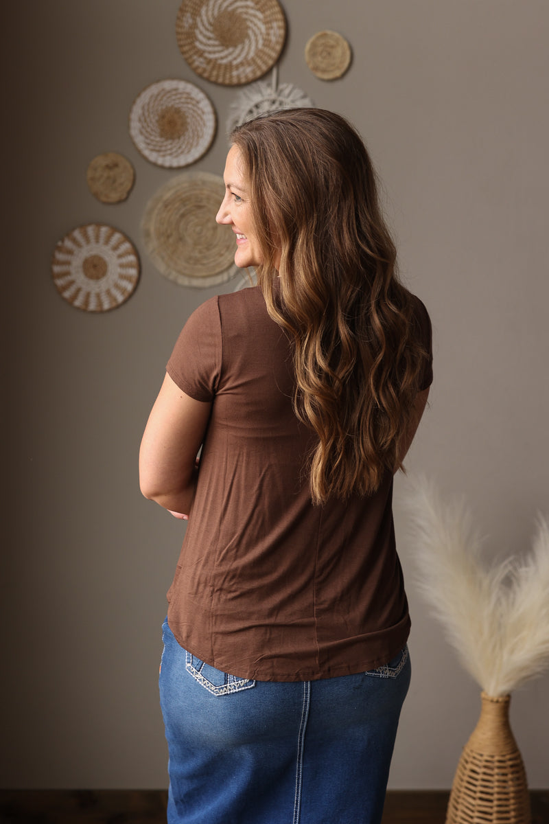 Woman wearing a brown shirt and blue jean skirt standing in front of decorative wall art.