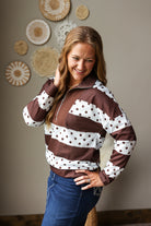 A woman wearing a brown and white striped quarter-zip sweatshirt with polka dot details on the sleeves and chest, paired with blue jean skirt, standing in front of a neutral wall decorated with circular woven wall art.