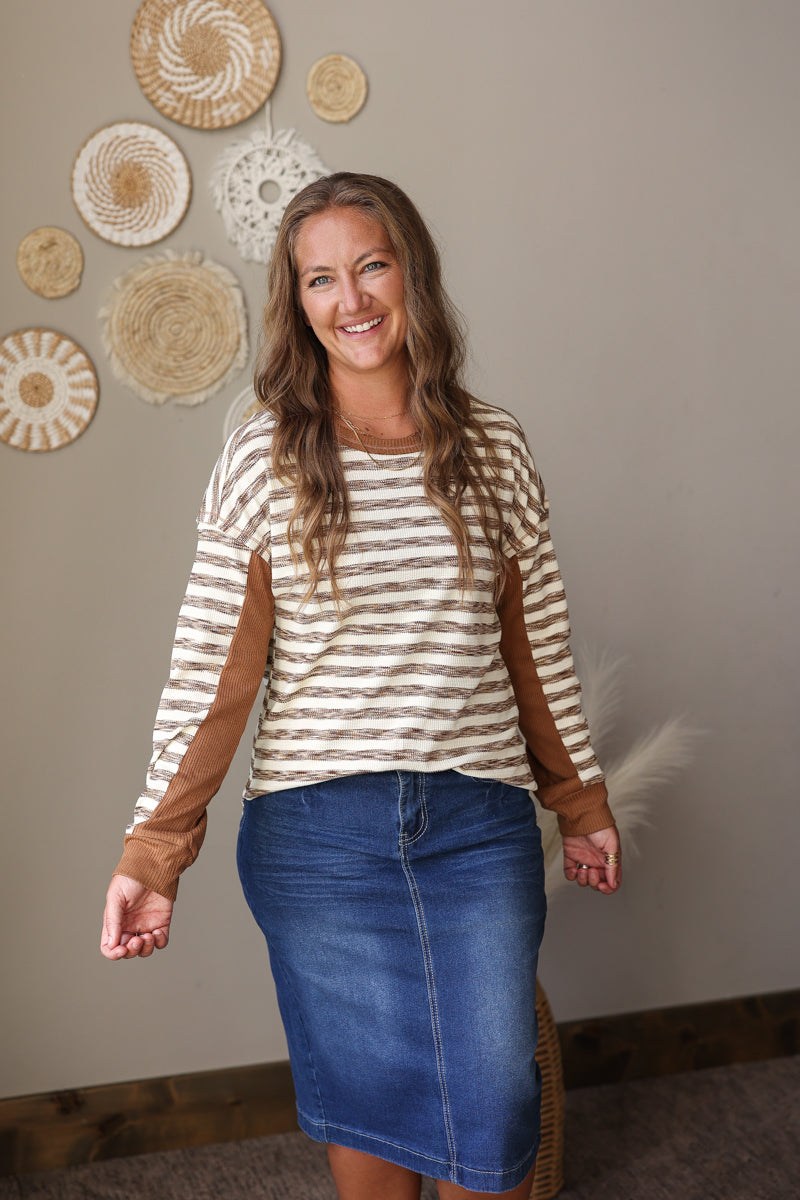 A woman modeling a long-sleeve striped top with brown elbow patches and a blue denim skirt, standing in front of a neutral wall decorated with woven wall art.