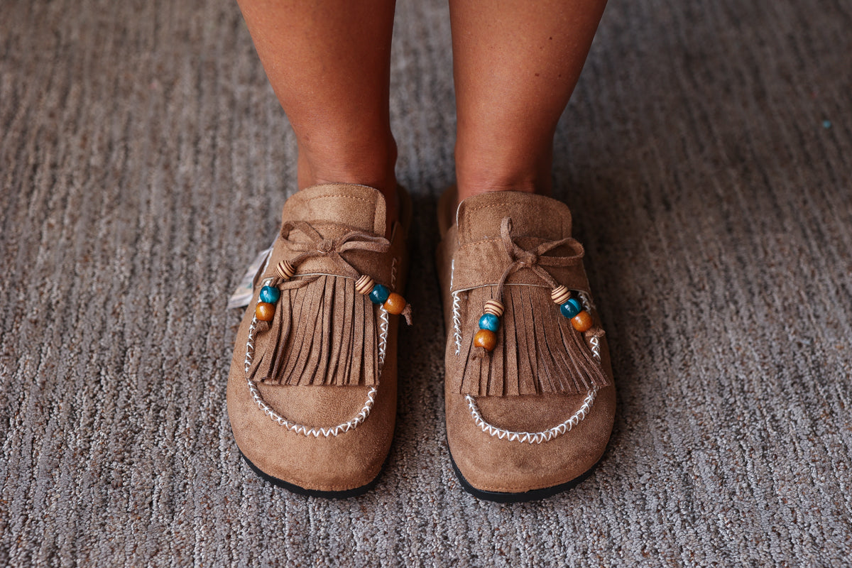 A pair of tan suede moccasin-style slip-on shoes with fringe, decorative beads, and white contrast stitching, shown being worn on a person's feet over a gray textured carpet.