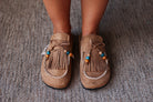 A pair of tan suede moccasin-style slip-on shoes with fringe, decorative beads, and white contrast stitching, shown being worn on a person's feet over a gray textured carpet.