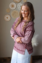 A woman with long light brown hair is wearing a dusty rose long-sleeve sweatshirt and light blue jean skirt, standing in a room with a neutral gray wall decorated with circular woven wall art.