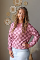 A smiling woman with long light brown hair stands indoors wearing a pink and mauve checkerboard long-sleeve top and a light blue skirt. The background shows a warm gray wall with decorative woven wall art and a vase with pampas grass.