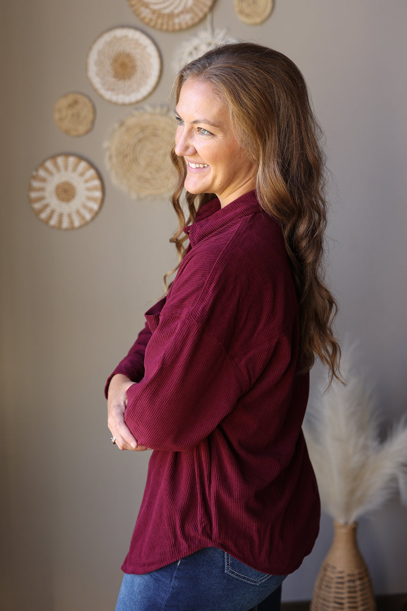 Side profile of a wine long sleeve corduroy shirt with a curved hem over a denim skirt.