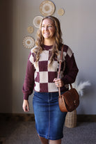 Angled front view of a brown checkered sweater with cream squares and a brown shoulder bag.