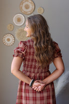 A woman with long wavy brown hair is shown from the back wearing a red and beige plaid dress. She is standing against a gray wall decorated with circular woven wall art.