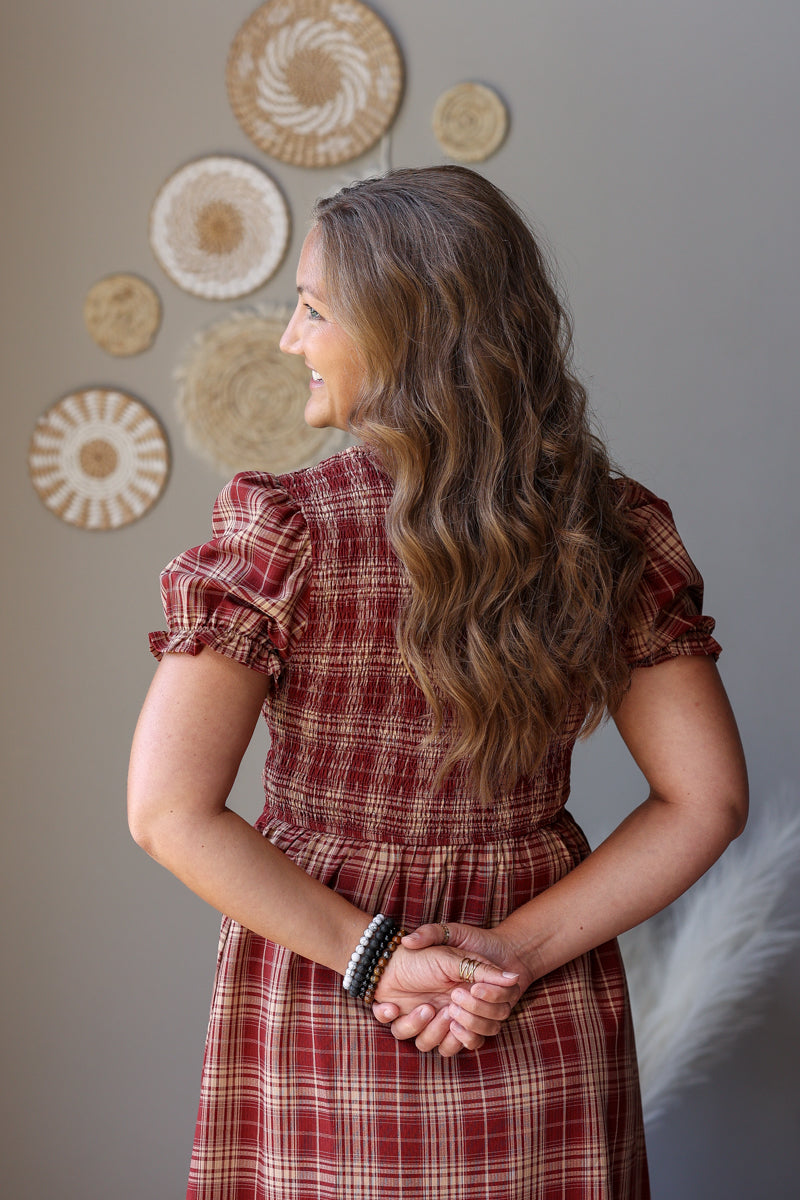 A woman with long wavy brown hair is shown from the back wearing a red and beige plaid dress. She is standing against a gray wall decorated with circular woven wall art.