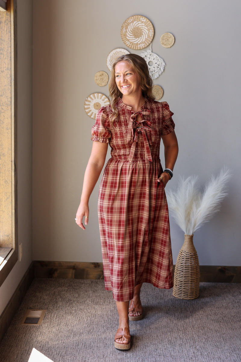 A woman modeling a short-sleeve red and brown plaid midi dress with a ruffled tie-neck, standing in a room with a beige wall decorated with circular wall art and a vase of pampas grass.