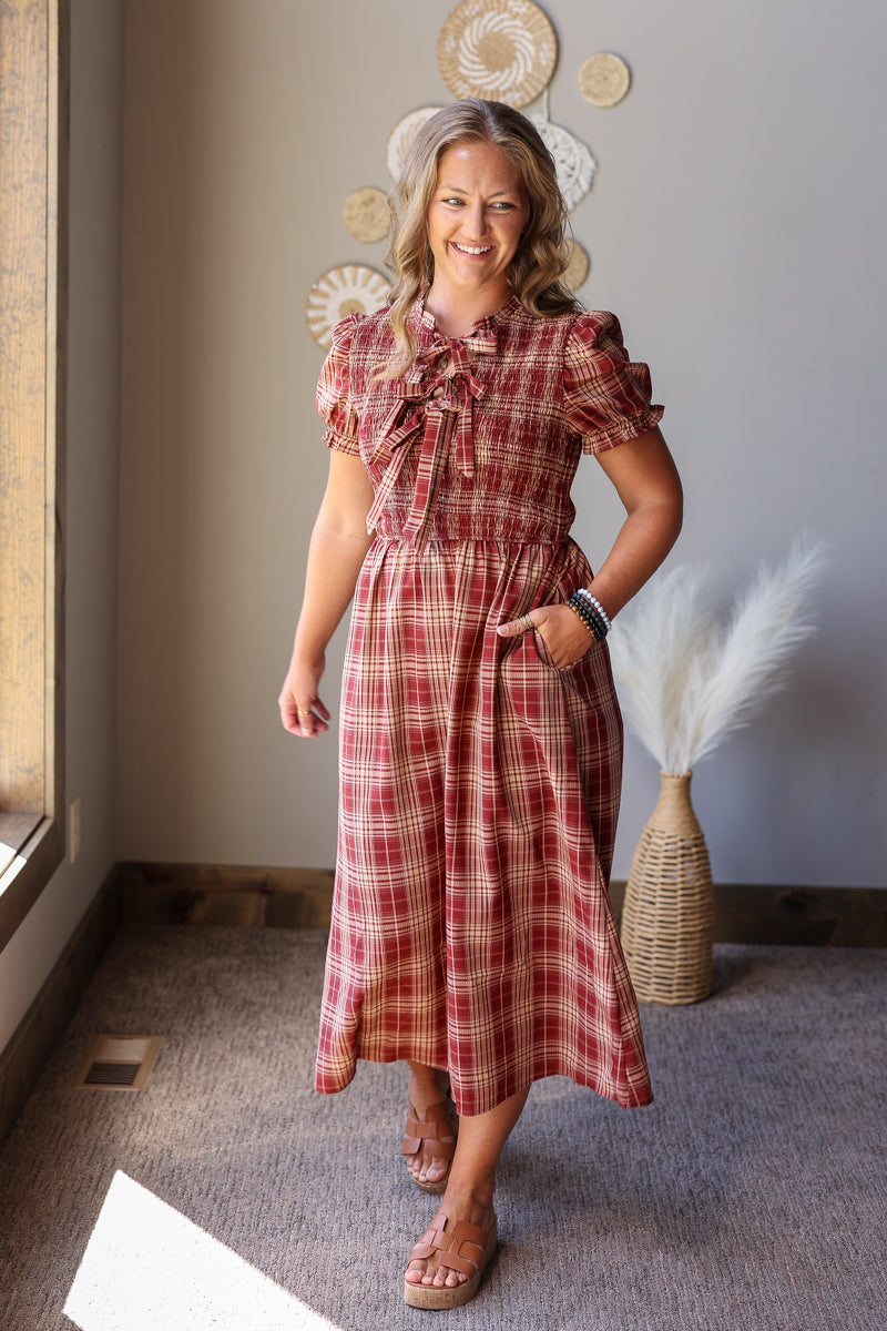 A woman modeling a red and brown plaid midi dress with short puff sleeves, a ruffled tie neckline, and side pockets, standing in a styled indoor room.