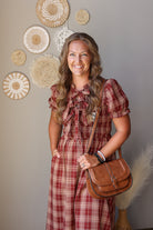 A woman with long wavy blonde hair stands indoors wearing a red and beige plaid midi dress with puff sleeves and a large bow at the neckline. She is holding a brown leather crossbody bag and smiling at the camera.