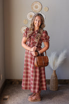 A woman stands indoors wearing a red and beige plaid midi dress with puff sleeves and holding a brown leather shoulder bag. The background shows a neutral wall with decorative woven wall art and a vase with dried pampas grass.