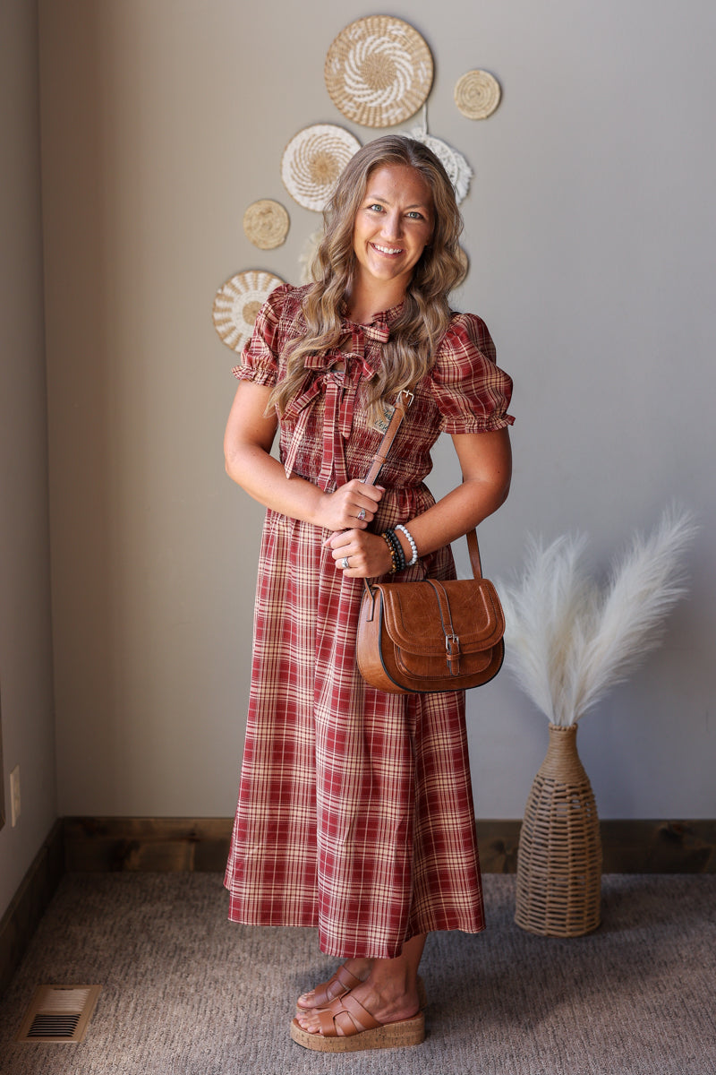 A woman stands indoors wearing a red and beige plaid midi dress with puff sleeves and holding a brown leather shoulder bag. The background shows a neutral wall with decorative woven wall art and a vase with dried pampas grass.