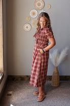 A woman modeling a red and brown plaid short-sleeve midi dress with a tie-neck detail, standing in a room with a gray wall decorated with circular wall art.