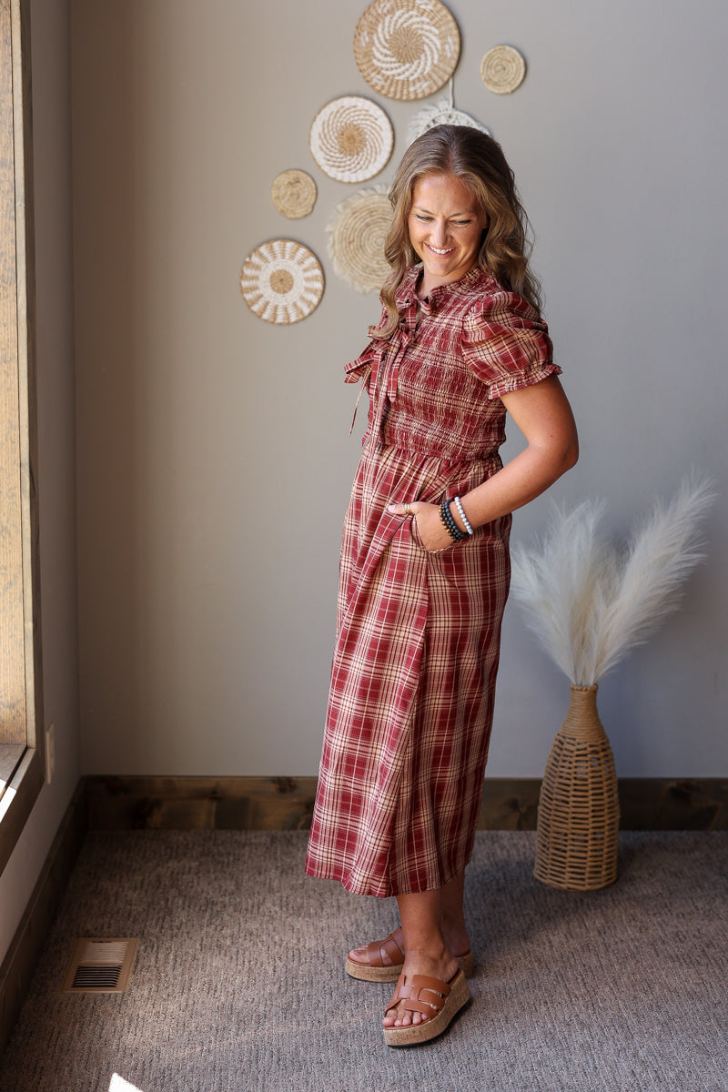 A woman modeling a red and brown plaid short-sleeve midi dress with a tie-neck detail, standing in a room with a gray wall decorated with circular wall art.