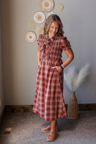 A woman modeling a red and white plaid midi dress with ruffled short sleeves and a tie-neck detail, standing in a styled room with a gray wall, decorative woven wall pieces, and a vase with pampas grass.