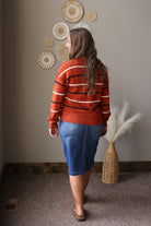 A woman is shown from behind wearing a rust-orange knit sweater with black and white horizontal stripes, paired with a blue denim skirt. She is standing in a room with a neutral gray wall decorated with circular woven wall art and a wicker vase with dried pampas grass.