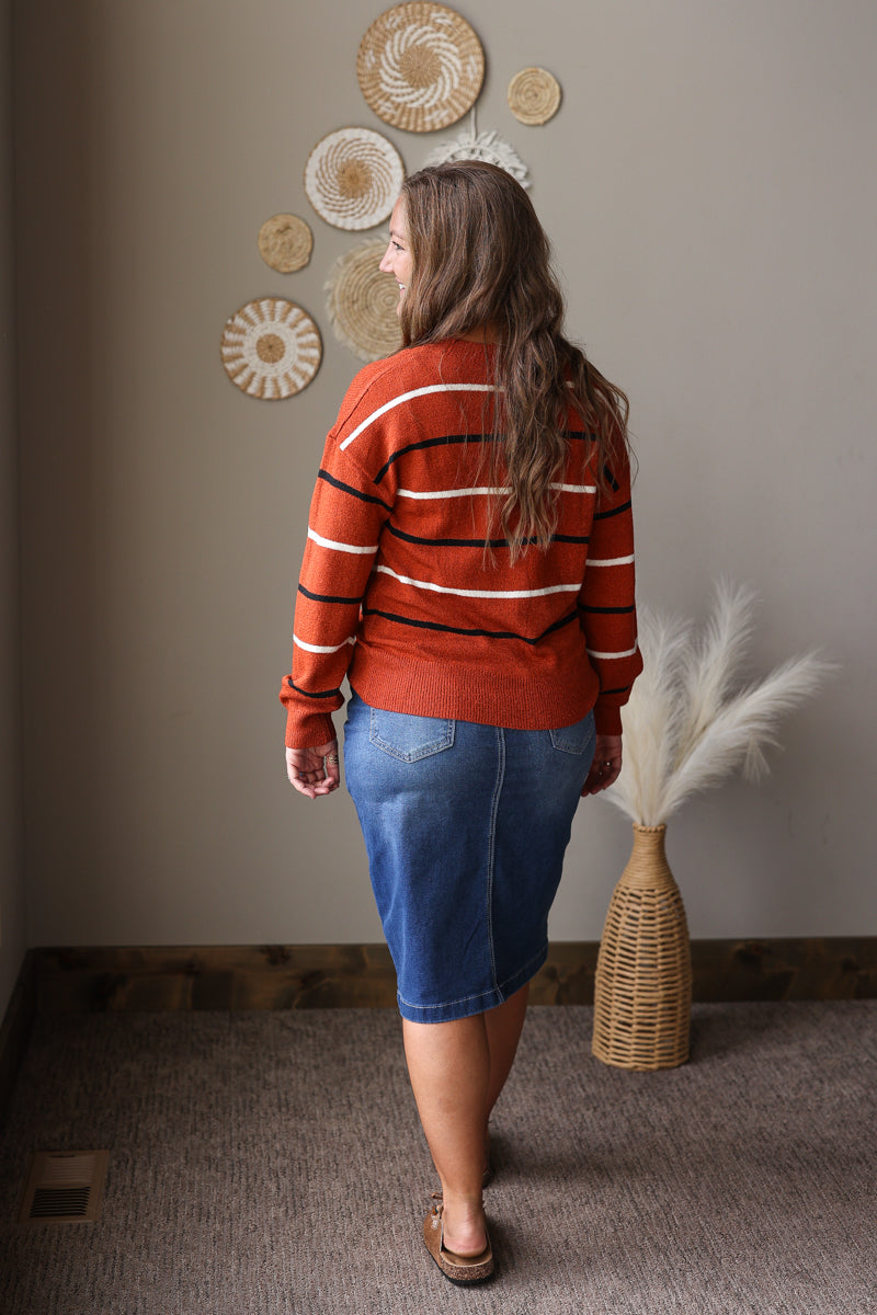 A woman is shown from behind wearing a rust-orange knit sweater with black and white horizontal stripes, paired with a blue denim skirt. She is standing in a room with a neutral gray wall decorated with circular woven wall art and a wicker vase with dried pampas grass.