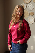 A woman with blonde hair smiling and looking to the side, wearing a dark red half-zip sweatshirt and blue jean skirt, standing in front of a wall decorated with circular woven wall art.