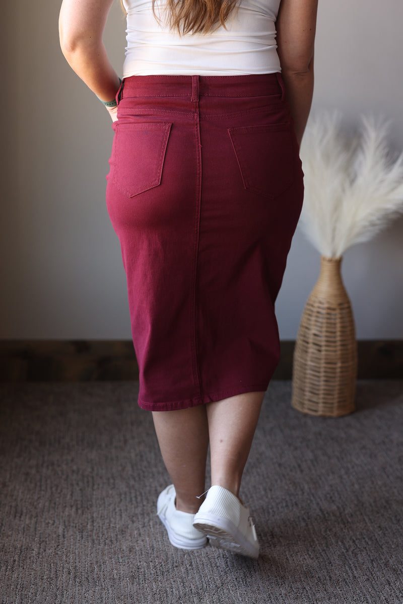 Person wearing a burgundy skirt with a white top and white shoes, standing in a room with a textured wall and wicker basket.