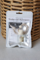 A car oil diffuser in a clear resealable pouch with a white label reading 'Teakwood Mahogany' and 'Honey Jar Creations,' placed in front of a woven wicker basket on a light surface.