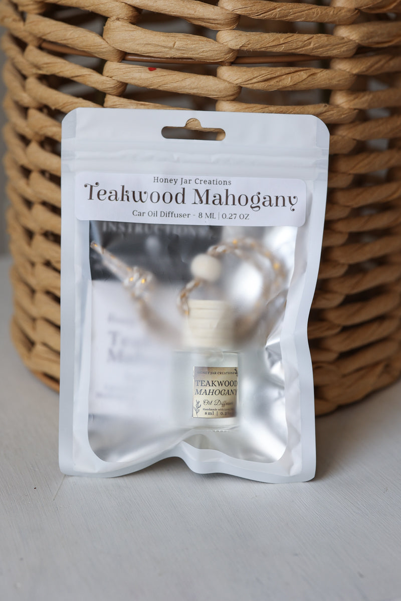 A car oil diffuser in a clear resealable pouch with a white label reading 'Teakwood Mahogany' and 'Honey Jar Creations,' placed in front of a woven wicker basket on a light surface.