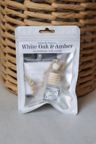 A car oil diffuser in a white resealable pouch labeled 'White Oak & Amber' by Honey Jar Creations, placed in front of a woven wicker basket on a light gray surface.