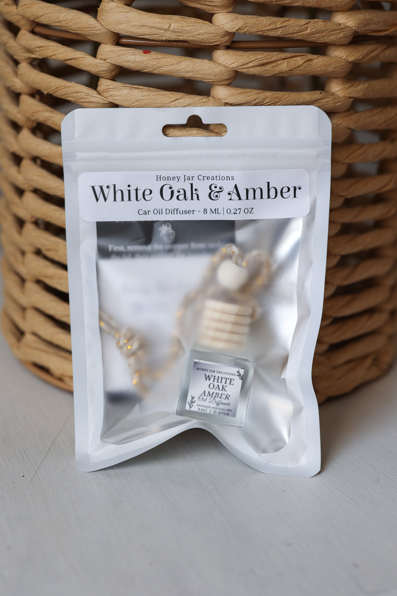 A car oil diffuser in a white resealable pouch labeled 'White Oak & Amber' by Honey Jar Creations, placed in front of a woven wicker basket on a light gray surface.