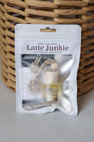 A Honey Jet Creations 'Latte Junkie' car oil diffuser in its white resealable packaging, displayed in front of a woven wicker basket on a light gray surface.