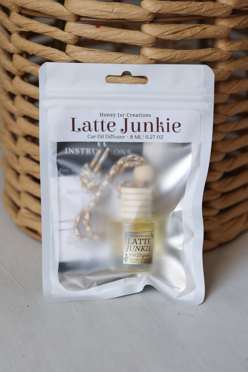 A Honey Jet Creations 'Latte Junkie' car oil diffuser in its white resealable packaging, displayed in front of a woven wicker basket on a light gray surface.