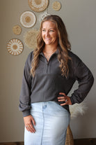 A woman with long brown hair smiling and posing in a dark gray ribbed quarter-zip pullover sweater and a light blue denim skirt, standing against a beige wall decorated with circular woven wall art.