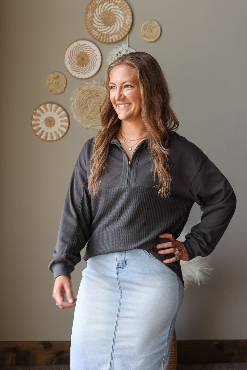 A woman wearing a dark gray quarter-zip long-sleeve top and a light blue denim skirt, standing in front of a beige wall decorated with circular woven wall art.