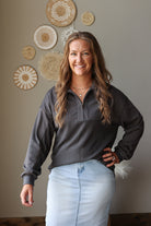 A woman smiling and posing indoors, wearing a dark charcoal gray ribbed quarter-zip long-sleeve pullover and a light blue denim skirt. She is standing in front of a beige wall decorated with circular woven wall art.