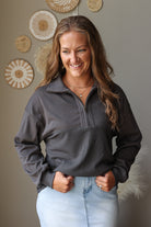 A woman smiling and wearing a dark gray quarter-zip pullover sweater with a textured waffle-knit lower front, paired with light blue jean skirt, standing against a neutral wall with decorative woven wall art.