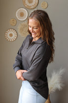 A woman wearing a dark gray quarter-zip long-sleeve top and light blue jean skirt, standing in a room with a neutral gray wall decorated with circular woven wall art.