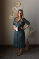 Woman in a grey dress holding a mug, standing in a room with decorative wall art.