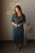 Woman in a grey dress standing in a room with decorative wall art and a vase.