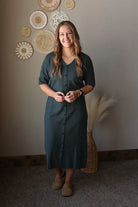 Woman in a grey dress standing in a room with decorative wall art and a plant.