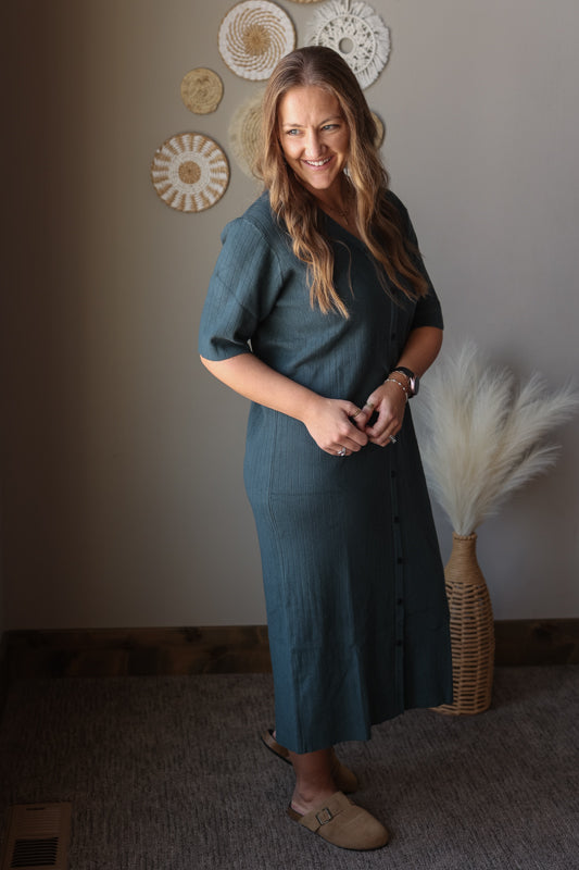 Woman in a gray dress standing in a room with decorative wall art and a vase of pampas grass.