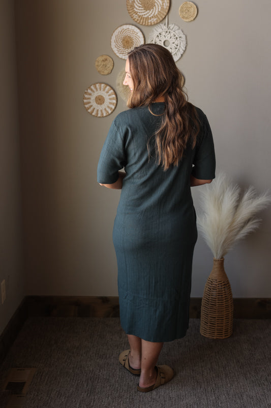 Woman in a gray dress standing in a room with decorative wall art and a vase.