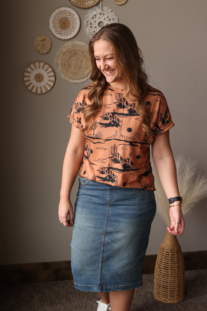 Woman wearing a patterned western tee and denim skirt standing indoors with decorative wall art in the background.